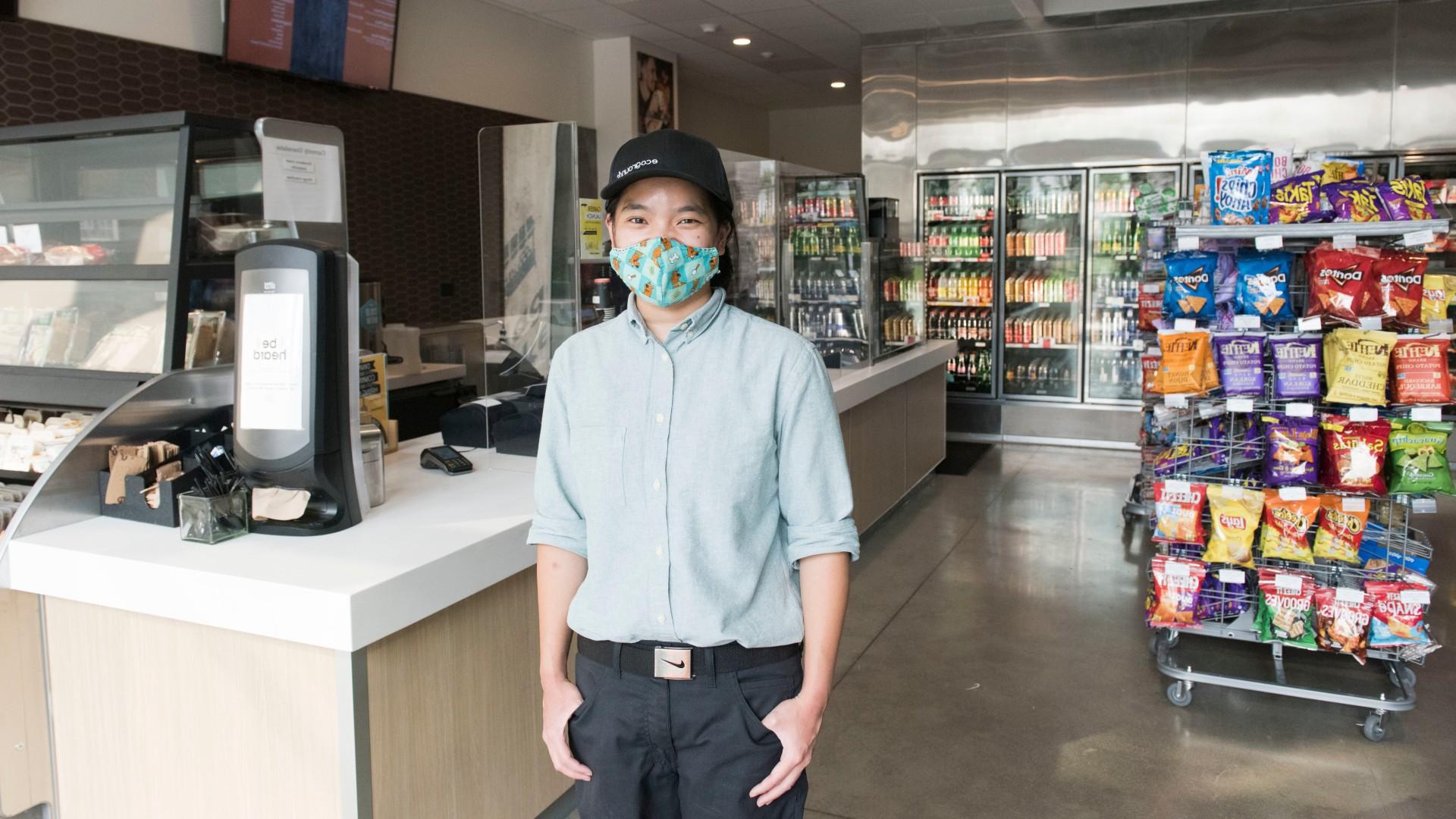 Person with a baseball cap and a facial covering poses for a photo in a convenience store.